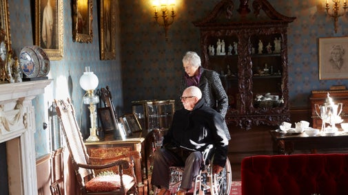 A lady pushes a man in a wheelchair as they look at photographs on a table in a room with lots of period collection items